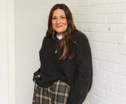 Azieta Amini stands against a white brick wall wearing a dark charcoal gray knit sweater and a green and beige plaid skirt.