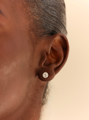 A close-up of a person's ear wearing a small round stud earring with a clear gemstone in a silver-toned setting, against a plain light beige background.