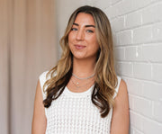 Athena Ambrogi wearing layered white gold necklaces, leaning against a white brick wall.