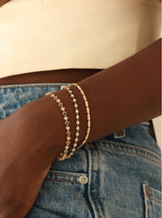 Three delicate gold bracelets stacked on a person's wrist, shown with blue denim jeans and a cream-colored top.