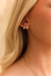 A close-up of a model's ear wearing a small gold huggie hoop earring with a tiny clear gemstone, next to blonde hair.