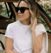 Woman wearing sunglasses, a white shirt, and a swan necklaces sitting inside a vehicle.