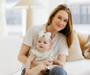Kat Heckman is sitting on a white sofa in a bright living room, holding her baby Vivi.
