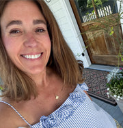 Woman wearing a shall charm,smiling outdoors with a wooden door and plants in the background.