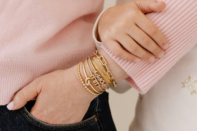 Close-up of a hand wearing multiple gold bracelets on a pink sweater background