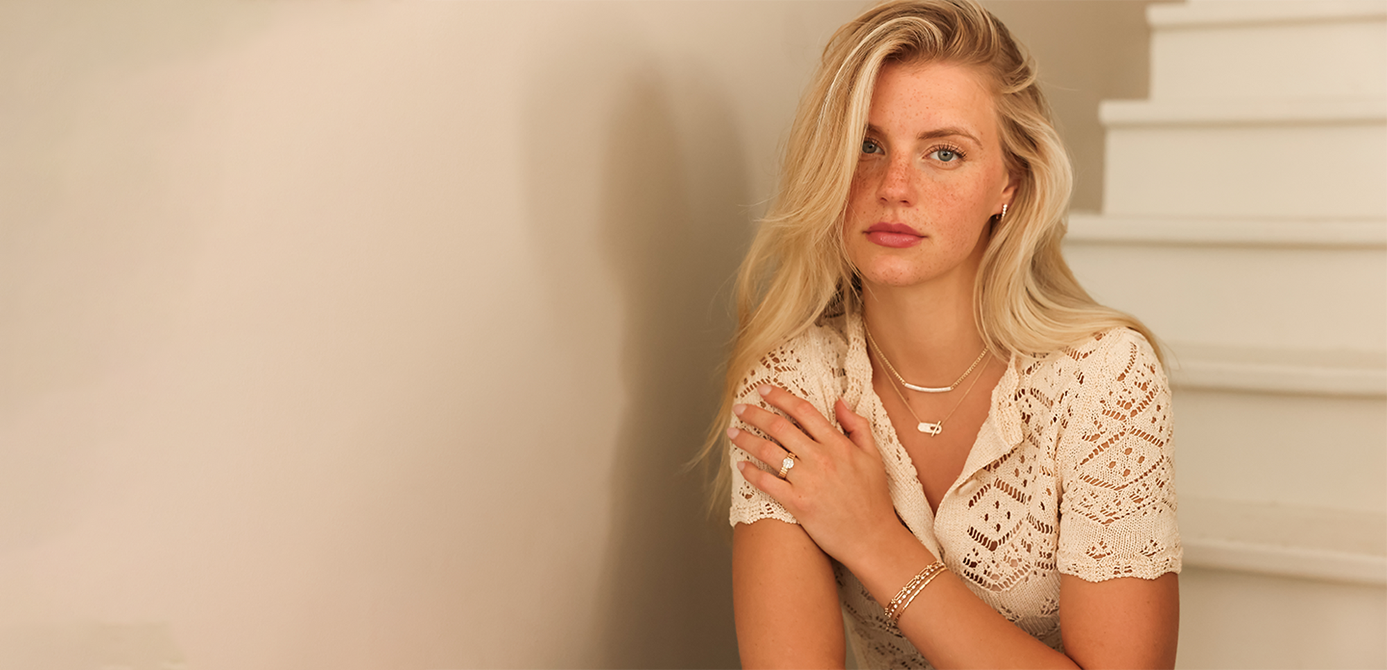 Woman in a white lace top standing against a neutral wall.