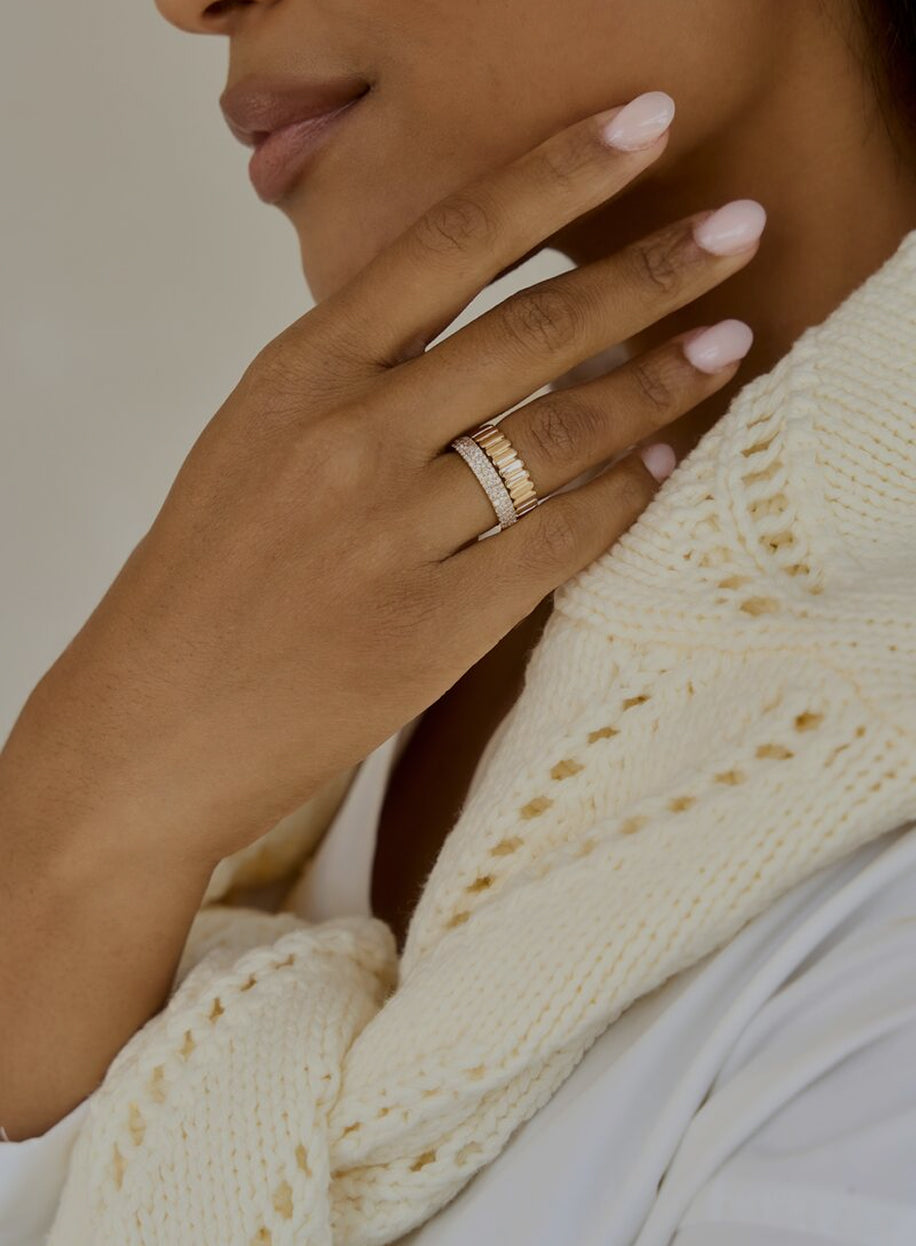 Close-up of a hand wearing a gold ring with a textured band, and a thick pave band, against a neutral background.